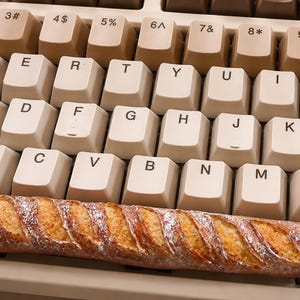 May include: A long, golden-brown baguette with a scored, flour-dusted crust resting horizontally across the keys of a beige computer keyboard. The keyboard features standard QWERTY layout keys with dark brown lettering on light beige square caps.