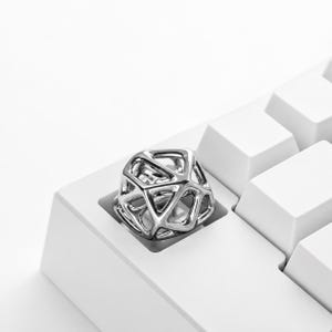 Puede incluir: Una tecla de teclado metálica de color plateado con un intrincado diseño de celosía geométrica de marco abierto. La tecla tiene forma de poliedro multifacético con aberturas triangulares y pentagonales huecas sobre un teclado blanco.