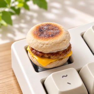 以下が含まれることがあります： 小さな朝食サンドイッチの形をしたカスタムメカニカルキーボードのキーキャップ。キーキャップは、トーストされたイングリッシュマフィンの上部、茶色のソーセージパティ、黄色いチーズのスライス、そしてマフィンの下部で構成されており、白いキーボードに取り付けられています。