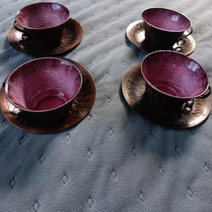 May include: Four tea cups and matching saucers featuring a deep purple, textured interior finish and a dark metallic exterior. Each saucer displays a decorative, embossed leaf-like pattern around the rim, complementing the dark metal cups.