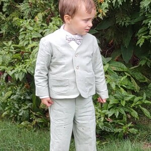 May include: A young boy wearing a light green suit with a bow tie. He is standing in a grassy area with green bushes behind him.
