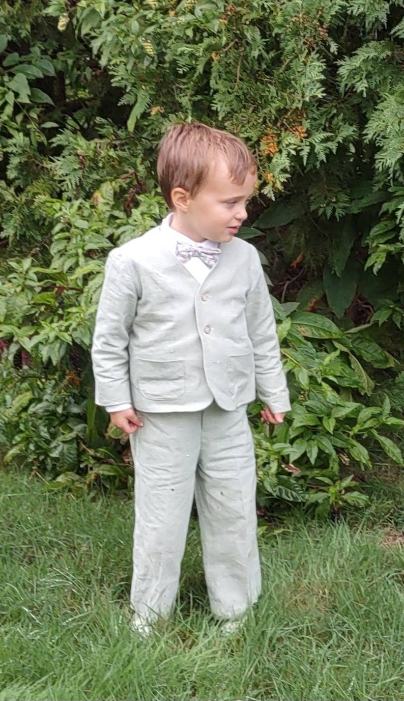 May include: A young boy wearing a light green suit with a bow tie. He is standing in a grassy area with green bushes behind him.