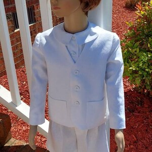 May include: A white linen suit for a young boy. The suit includes a jacket with a peter pan collar and a pair of shorts. The jacket has a button-up front and two pockets. The shorts have a button-up front and a pleated waistband.