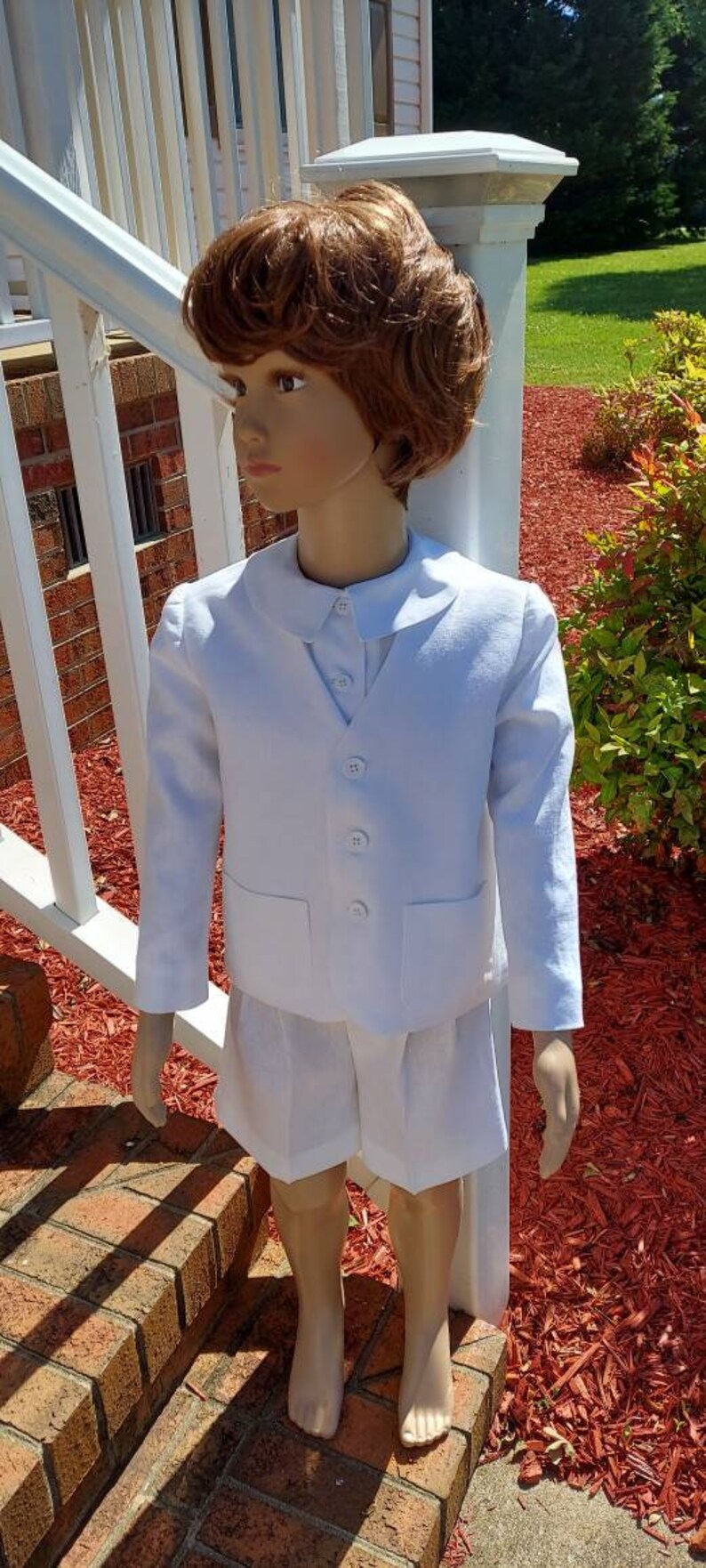 May include: A white linen suit for a young boy. The suit includes a jacket with a peter pan collar and a pair of shorts. The jacket has a button-up front and two pockets. The shorts have a button-up front and a pleated waistband.
