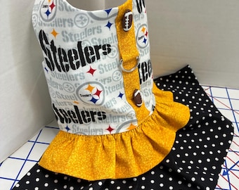 May include: A pet harness dress featuring a white fabric with a repeating Pittsburgh Steelers logo and text pattern. It has a mustard yellow ruffled waist, a vertical yellow strip with two football-shaped buttons, and a black skirt with white polka dots.