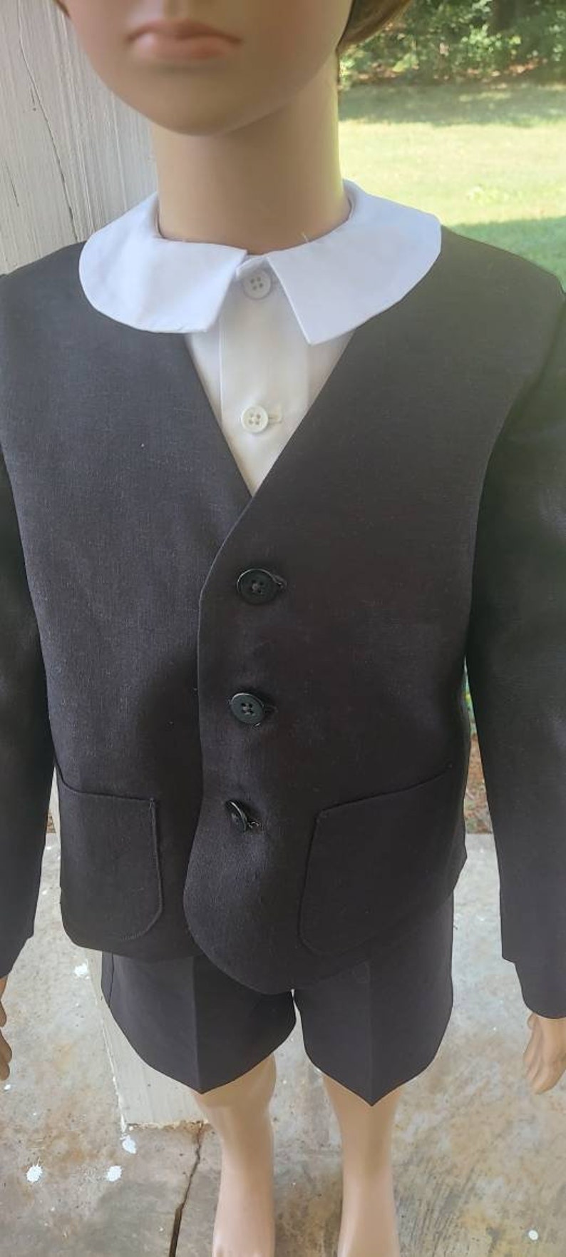 May include: A black suit with a white collared shirt. The suit has a vest with four buttons and two pockets. The suit is worn by a child.