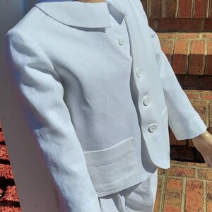 May include: A white linen suit with a peter pan collar and short pants. The suit has a single button closure and a pocket on the front. The suit is on a child-sized mannequin.