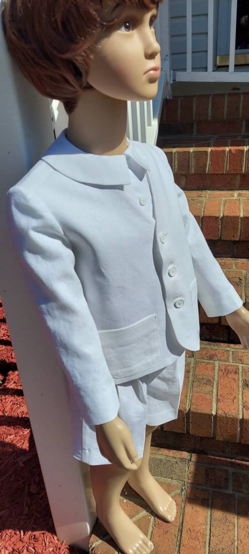 May include: A white linen suit with a peter pan collar and short pants. The suit has a single button closure and a pocket on the front. The suit is on a child-sized mannequin.