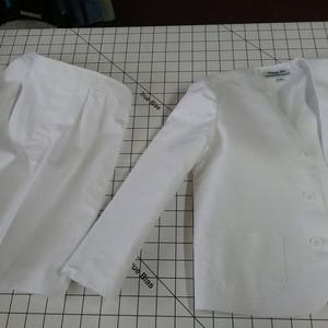 May include: A white skirt and blazer set. The blazer has a single button closure and a pocket on the front. The skirt is pleated and has a simple design.