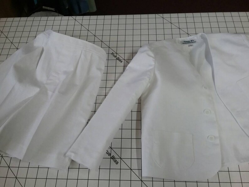 May include: A white skirt and blazer set. The blazer has a single button closure and a pocket on the front. The skirt is pleated and has a simple design.