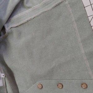 May include: A light green linen garment with three brown buttons. The garment has a seam down the center and a folded edge.