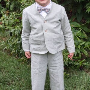 May include: A young boy wearing a light green linen suit with a patterned bow tie. He is standing in a grassy area and smiling.
