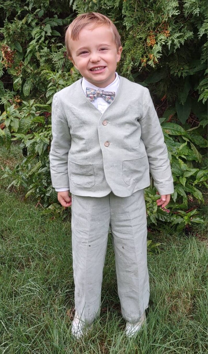May include: A young boy wearing a light green linen suit with a patterned bow tie. He is standing in a grassy area and smiling.