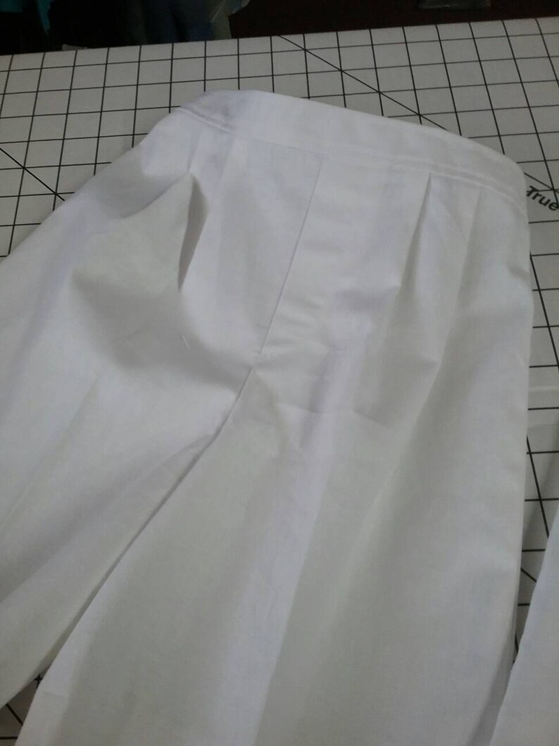 May include: White pleated pants with a single front pocket. The pants are made of a lightweight fabric and have a relaxed fit.