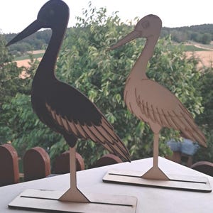 Könnte beinhalten: Zwei stehende Vogelfiguren aus flachem Material, die jeweils auf einem rechteckigen Sockel montiert sind. Ein Vogel ist einfarbig schwarz mit sichtbaren Federdetails, während der andere aus hellem Holz mit eingravierten Federumrissen besteht.