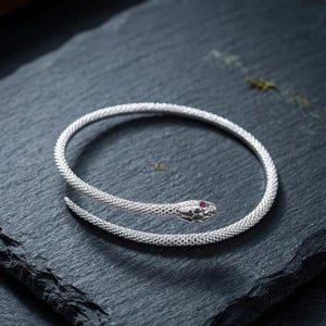 May include: A silver-toned wrap bracelet shaped like a coiled snake with a textured, scale-like surface. The snake head features a small, round, deep red gemstone set as an eye. The bracelet has an open, flexible design that curves into a circular shape.