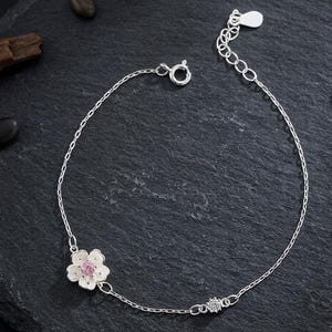 Peut inclure: Un bracelet à chaîne en argent orné d'une breloque en forme de fleur à cinq pétales avec une pierre centrale rose et une petite pierre transparente ronde. Le bracelet comprend une fine chaîne à maillons, un fermoir à ressort et une chaînette.