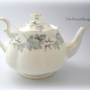 Pode incluir: Bule de porcelana creme com tampa, asa e bico. O bule é decorado com um padrão de folhas cinzentas e verdes e detalhes dourados. O texto "The French Budgie" é visível.