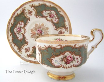 Peut inclure: Un service à thé vintage composé d'une tasse et d'une soucoupe. La tasse et la soucoupe sont blanches avec un motif floral vert et or. La tasse a une anse et une base dorées. La soucoupe a un bord festonné. Le texte "The French Budgie" est en bas.