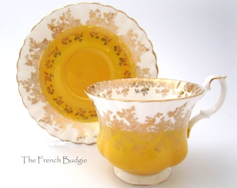 May include: A yellow teacup and saucer set with a white background and gold accents. The teacup and saucer have a delicate floral pattern.
