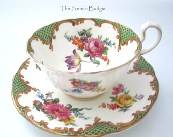 Può includere: Una tazza da tè e un piattino bianchi con un motivo floreale verde e oro. La tazza e il piattino hanno un bordo ondulato e un motivo floreale con fiori rosa, gialli e viola.