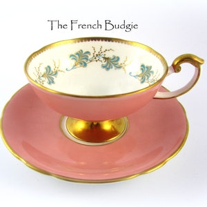 May include: A pink teacup and saucer set with a gold trim and a delicate blue and gold floral design. The teacup has a footed base.