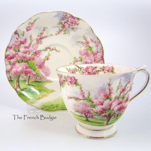 May include: A vintage teacup and saucer set. The white porcelain cup and saucer feature a pink cherry blossom design with gold trim. The cup has a pedestal base and a curved handle. The saucer has a scalloped edge.