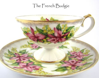 Puede incluir: Una taza de té y un platillo blancos con un diseño floral. La taza y el platillo presentan flores rosas y amarillas con hojas verdes sobre un fondo blanco. La taza tiene un borde dorado y el platillo tiene un borde dorado y una banda dorada alrededor del borde.
