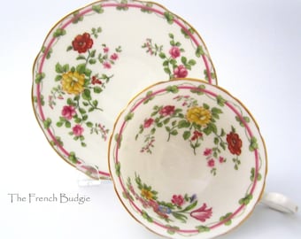 Può includere: Una tazza da tè e un piattino bianchi con un motivo floreale in tonalità rosa, giallo e rosso. Il motivo presenta un delicato disegno floreale con foglie verdi e un bordo dorato.