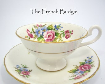 Puede incluir: Una taza de té y un platillo blancos con un borde dorado y un patrón floral. Las flores son rosas, azules y verdes.