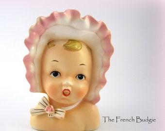 May include: A vintage porcelain baby doll head planter with a pink bonnet and a white bow with a pink rose. The doll has a painted face with closed eyes and a pink mouth.
