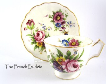 Puede incluir: Una taza de té y un platillo blancos con un patrón floral. La taza y el platillo tienen un borde dorado. Las flores son rosas, amarillas, azules y moradas.