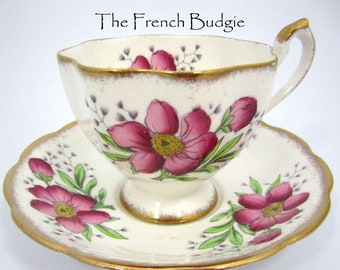 Puede incluir: Un juego de taza y platillo de té vintage con un diseño floral. La taza y el platillo son blancos con un borde dorado y presentan flores rosas con hojas verdes y tallos morados.