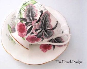 Puede incluir: Una taza de té y un platillo blancos con un diseño floral con orquídeas rosas y negras. La taza y el platillo tienen un borde dorado.