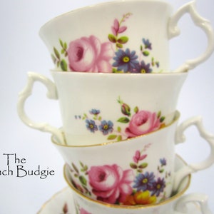 May include: A stack of four white teacups with floral designs. The cups have gold trim and feature pink roses, blue forget-me-nots, and green leaves.