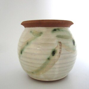 May include: A small, round ceramic vase with a cream-coloured body and a brown rim. The vase features green and tan abstract painted designs. The exterior has horizontal lines. The vase is suitable for holding small flowers or decorative items.