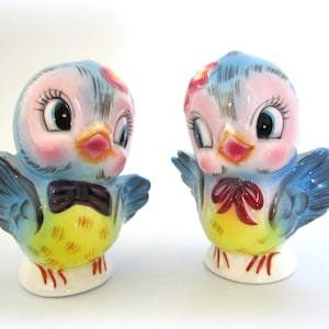 Pode incluir: Um par de saleiros e pimenteiros de cerâmica azul e amarelo em forma de pássaro com olhos grandes e detalhes pintados. Os pássaros usam laços e têm um visual caprichoso.