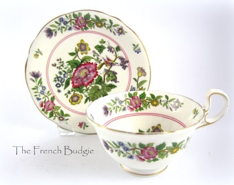 Puede incluir: Un juego de taza y platillo de té vintage con un patrón floral. La taza y el platillo son blancos con un borde dorado y presentan un diseño floral rosa, amarillo y verde con un pequeño pájaro.