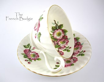 May include: A white teacup and saucer with a floral pattern of pink roses and green leaves. The cup and saucer have a gold trim.