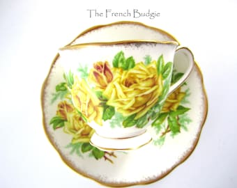 Può includere: Una tazza da tè e un piattino bianchi con un bordo dorato. La tazza da tè e il piattino sono decorati con un motivo floreale di rose gialle e foglie verdi.