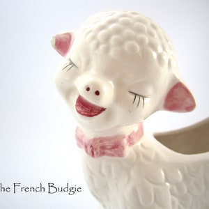 May include: A white ceramic planter shaped like a lamb with pink ears and a pink bow. The lamb has closed eyes and a smiling mouth.
