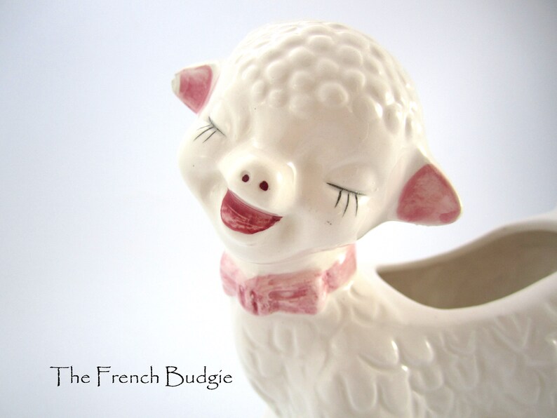 May include: A white ceramic planter shaped like a lamb with pink ears and a pink bow. The lamb has closed eyes and a smiling mouth.