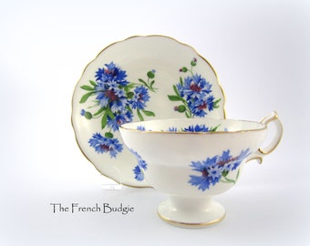 May include: A white teacup and saucer with a blue and green floral pattern. The floral pattern features blue cornflowers with green leaves and stems.