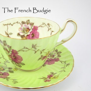 May include: A lime green teacup and saucer set with a floral design in pink, white, and green. The teacup and saucer have a gold trim. The text "The French Budgie" is at the top of the image.