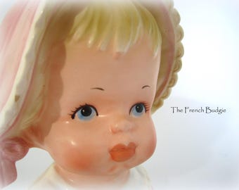 May include: A vintage porcelain doll planter with a pink and white hat. The doll has blonde hair, blue eyes, and a rosy complexion.