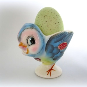 May include: A vintage ceramic egg cup shaped like a blue bird holding a green speckled egg. The bird has a pink face, orange beak, and black eyes. The base is white with red feet. A red sticker on the side reads "Andersen's Edition".