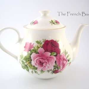 Może przedstawiać: Biały czajnik z kwiatowym wzorem z różowymi i czerwonymi różami. Czajnik ma pokrywkę i uchwyt.