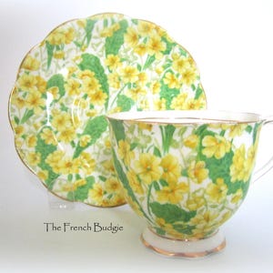 Può includere: Un set da tè con tazza e piattino con un motivo floreale. La tazza e il piattino sono bianchi con un design floreale giallo e verde. Il piattino ha un bordo ondulato.