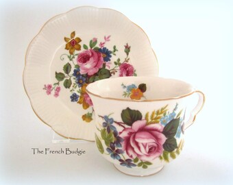 May include: A white teacup and saucer set with a floral pattern. The cup and saucer feature a pink rose, green leaves, and blue and yellow flowers. The saucer has a scalloped edge.