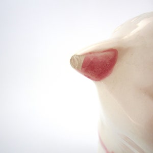 May include: Close-up of a white ceramic piggy bank with a pink ear and black painted details. The piggy bank is slightly chipped on the ear.